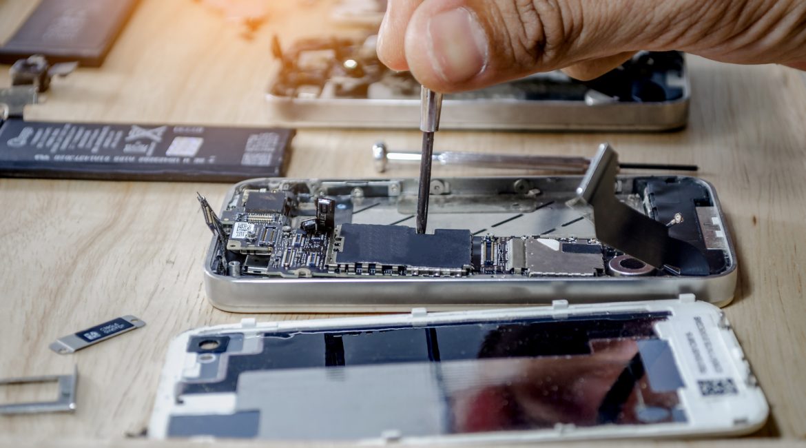 A technician's hand uses a small screwdriver to perform a repair on the intricate motherboard of a disassembled smartphone on a wooden workbench.