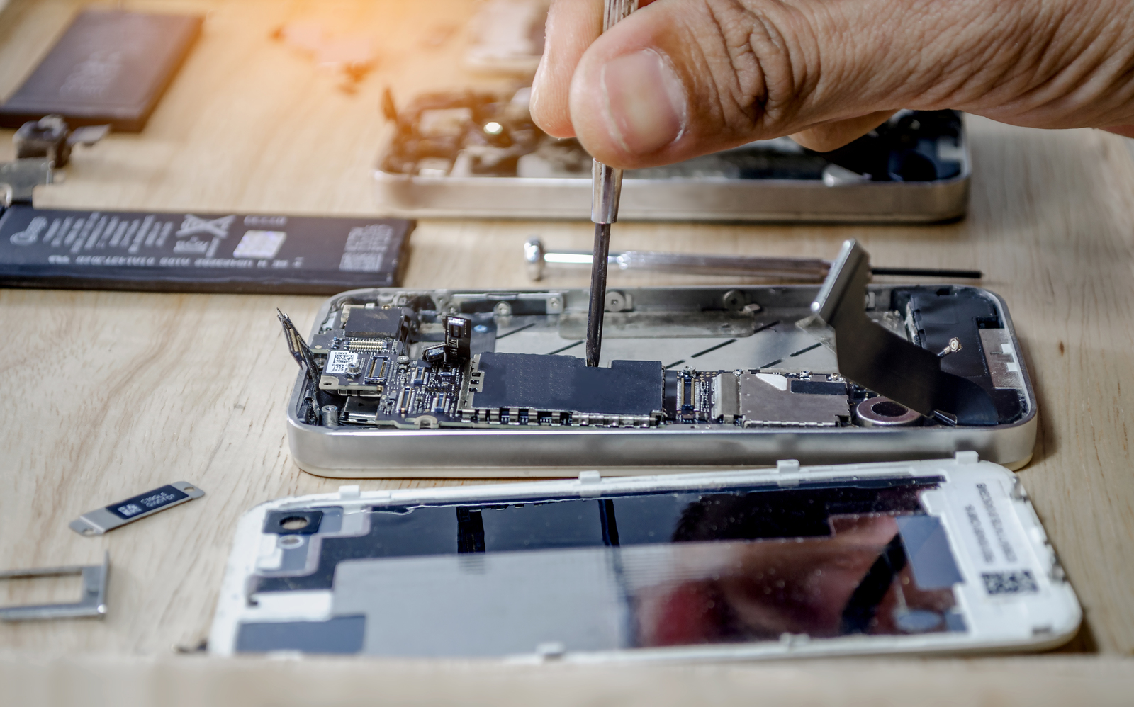 A technician's hand uses a small screwdriver to perform a repair on the intricate motherboard of a disassembled smartphone on a wooden workbench.