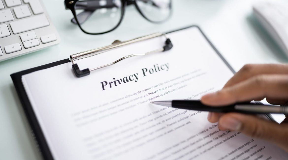 A person holds a pen over a privacy policy document on a clipboard, sitting on a clean desk with glasses and a keyboard.