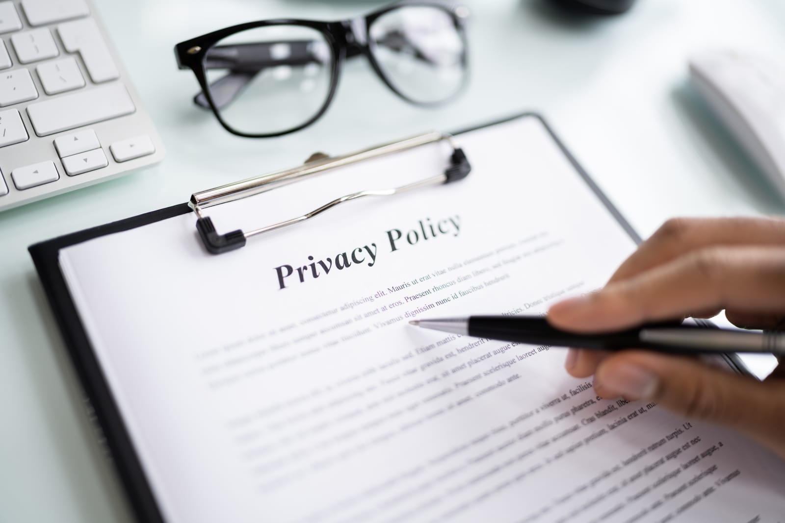 A person holds a pen over a privacy policy document on a clipboard, sitting on a clean desk with glasses and a keyboard.
