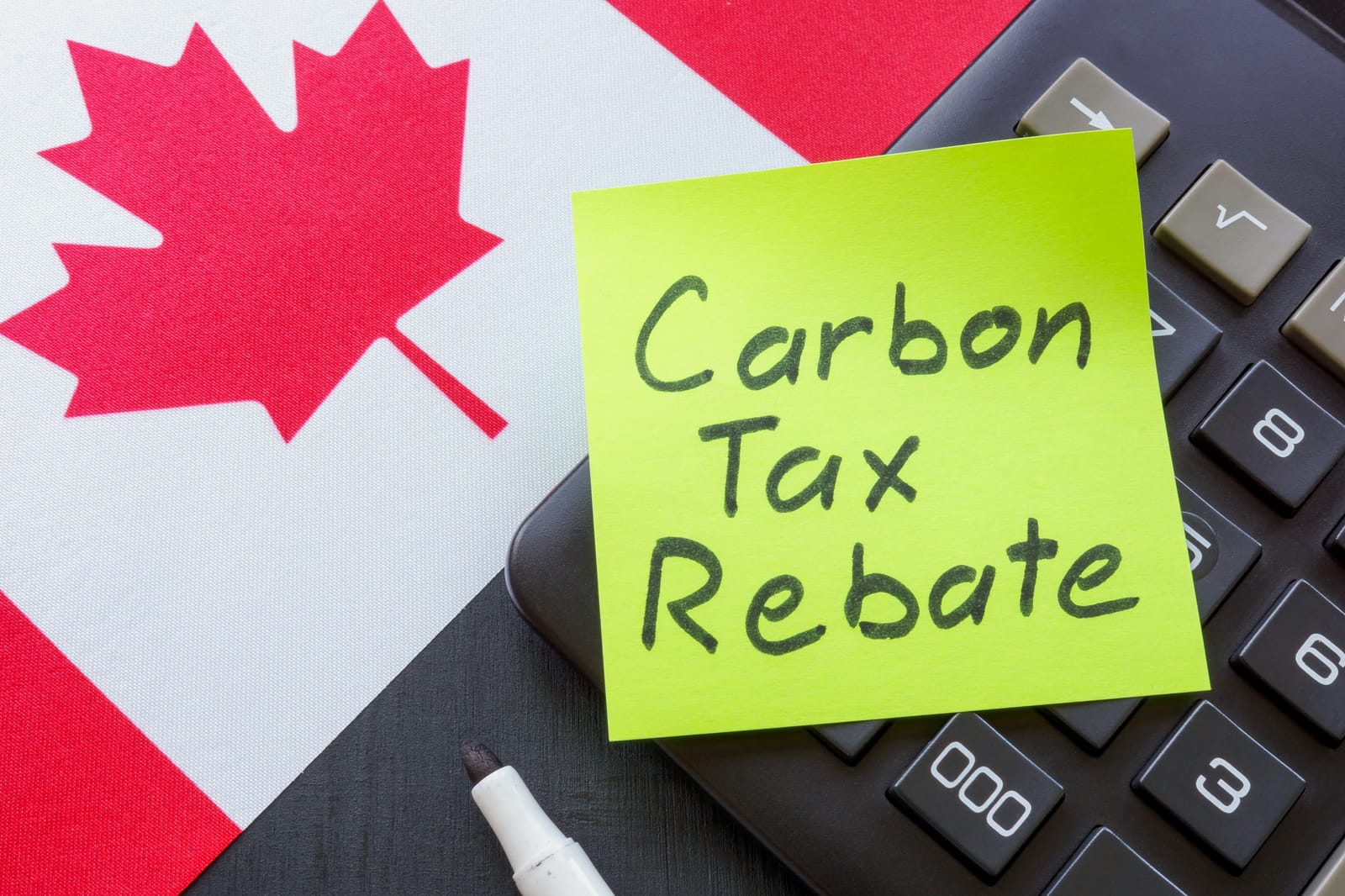 A sticky note with the words 'Carbon Tax Rebate' written on it sits on a calculator, which is placed on a Canadian flag, symbolizing Canada's financial rebate system for carbon pricing.