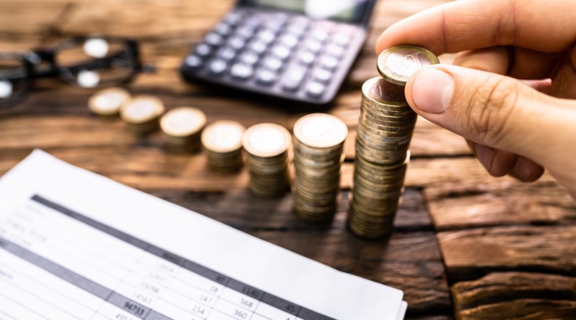 A person's hand stacking coins on a wooden desk next to a calculator and a financial spreadsheet, symbolizing the economic concepts of income and a basic income guarantee.