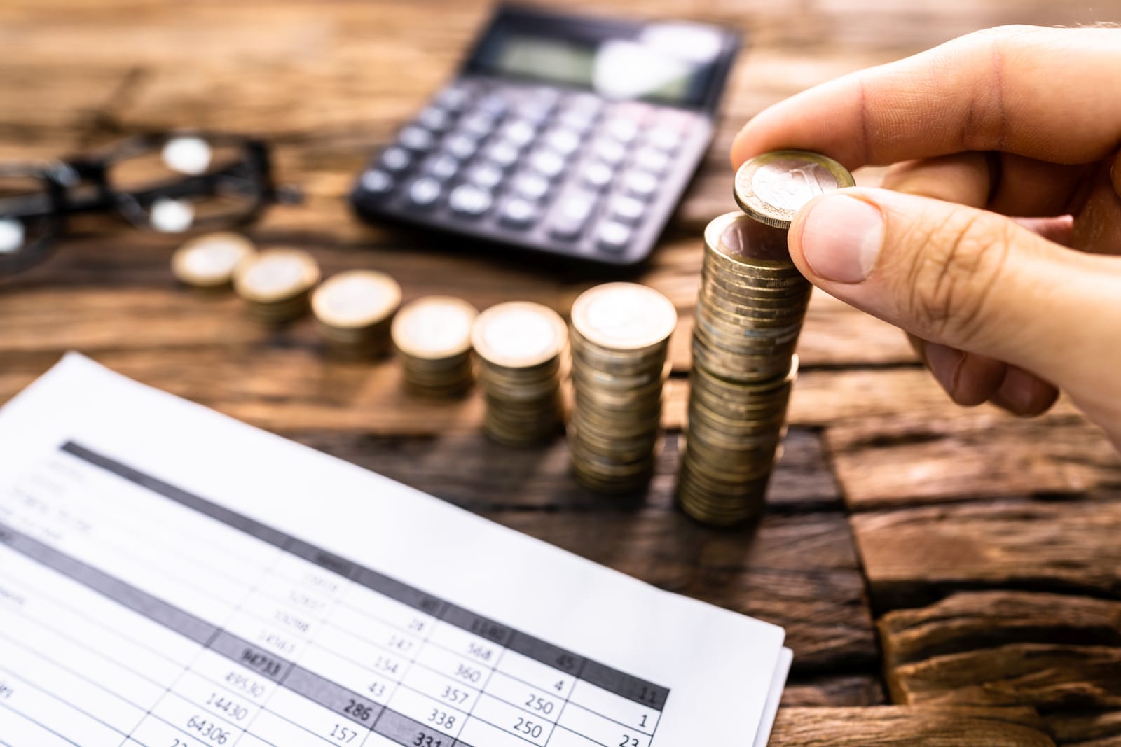 A person's hand stacking coins on a wooden desk next to a calculator and a financial spreadsheet, symbolizing the economic concepts of income and a basic income guarantee.