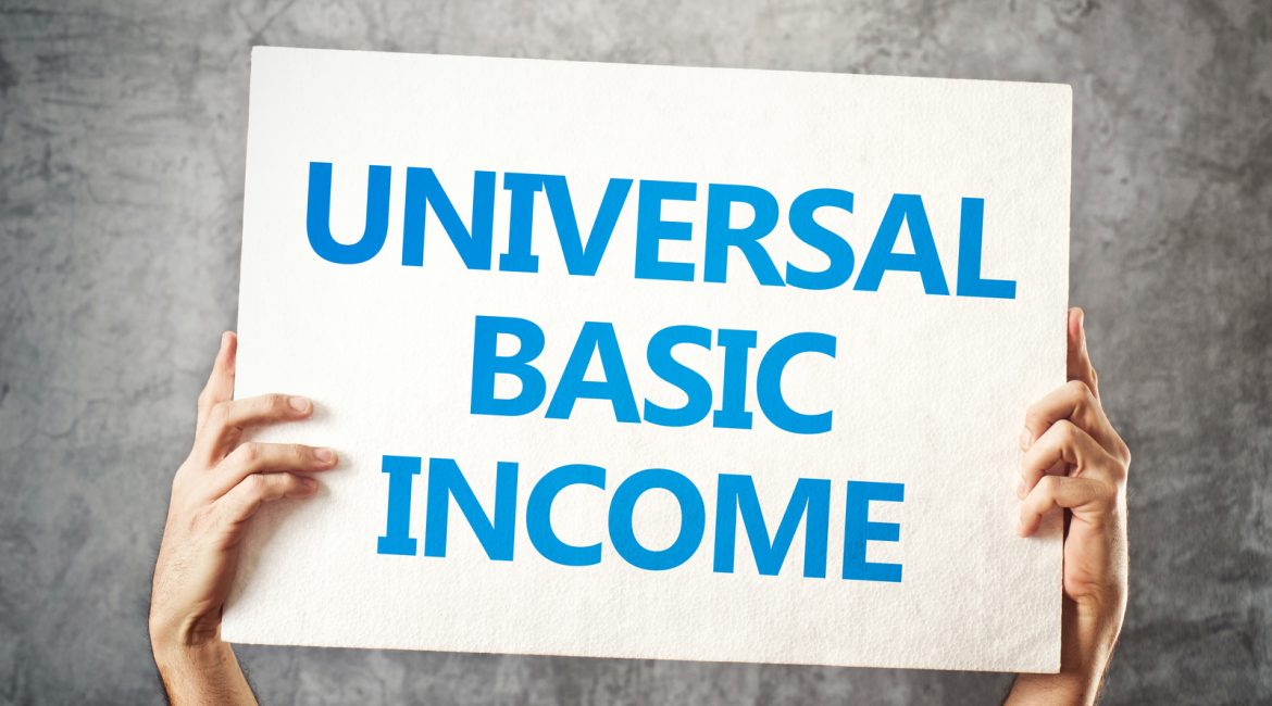 A person holds up a white sign with the blue words 'Universal Basic Income' against a grey textured wall.