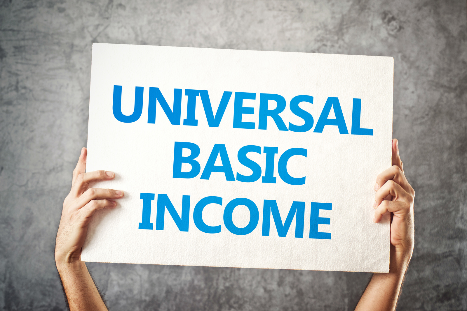 A person holds up a white sign with the blue words 'Universal Basic Income' against a grey textured wall.