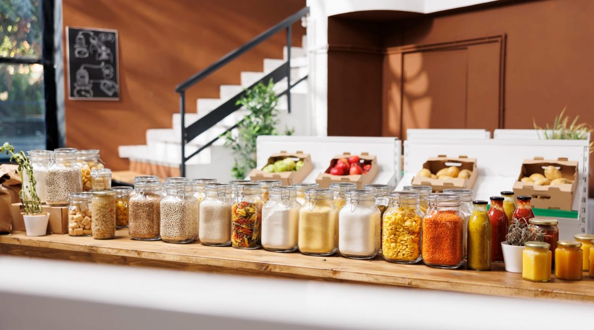 A long wooden counter in a bright, modern zero-waste store, lined with glass jars of bulk foods like grains and pasta, with fresh produce in the background