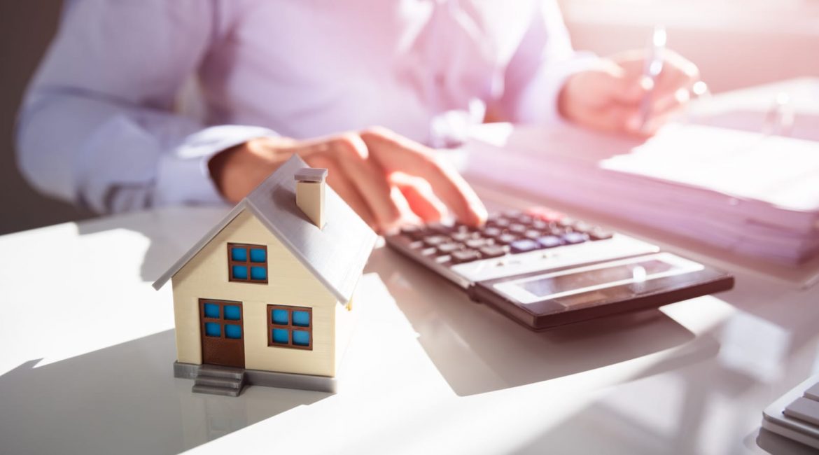 A person uses a calculator at a desk with a model of a house in the foreground, symbolizing the process of calculating rising home insurance costs.