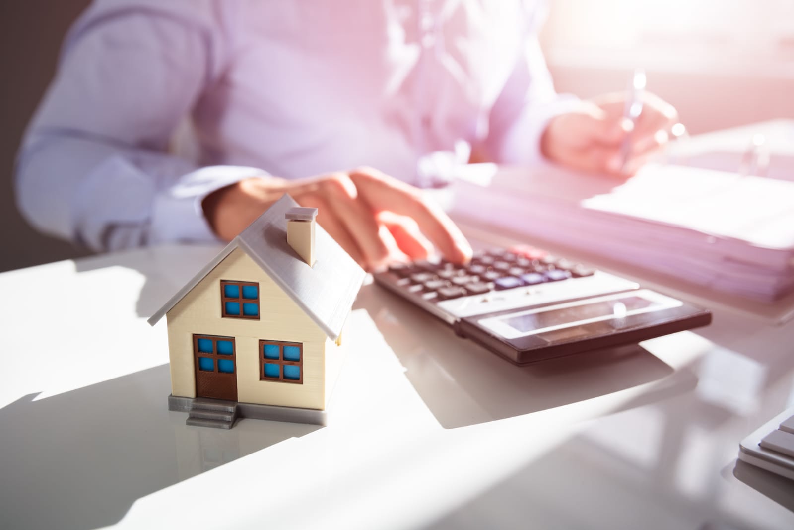 A person uses a calculator at a desk with a model of a house in the foreground, symbolizing the process of calculating rising home insurance costs.