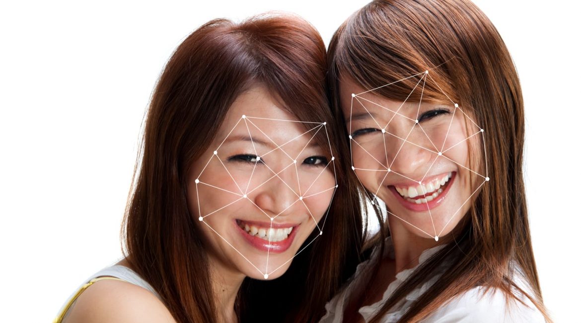Two smiling East Asian women have their faces scanned by a digital facial recognition overlay, representing the use of biometric identification technology.