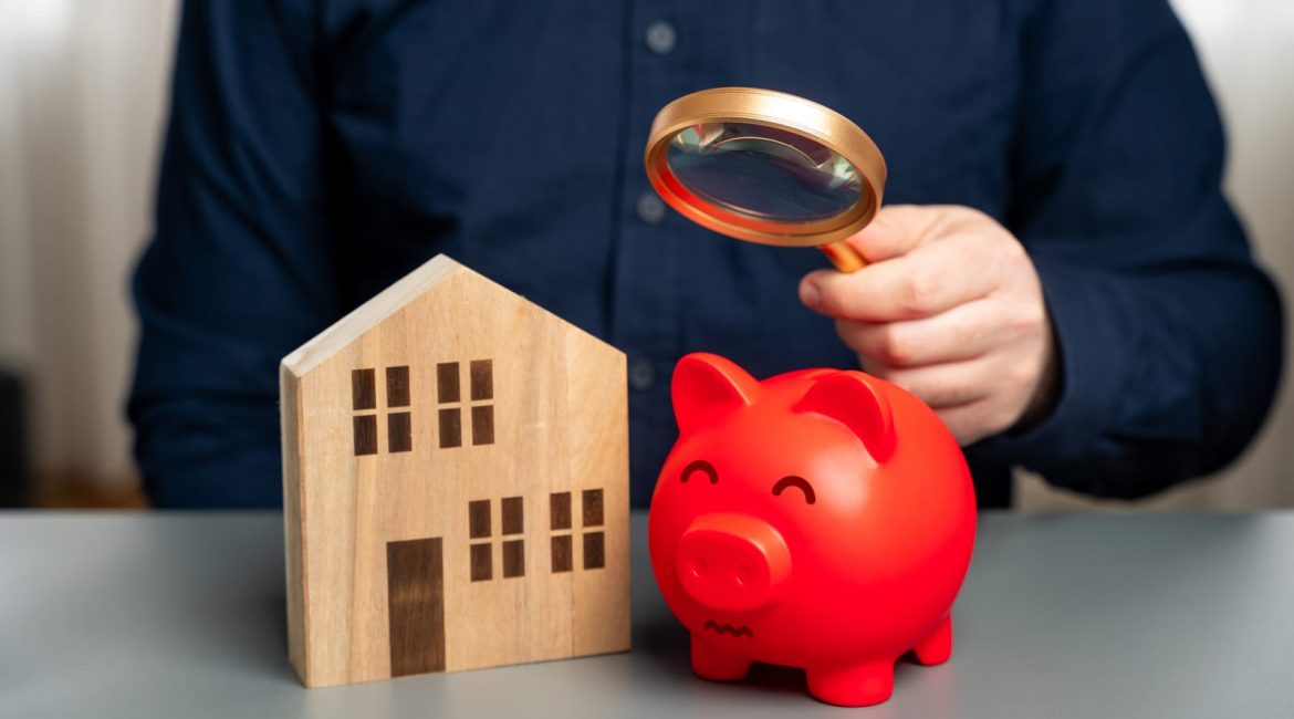 A person holds a magnifying glass over a red piggy bank that sits next to a wooden model of a house, symbolizing the financial scrutiny of housing affordability.