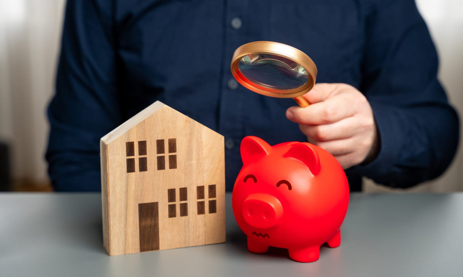A person holds a magnifying glass over a red piggy bank that sits next to a wooden model of a house, symbolizing the financial scrutiny of housing affordability.