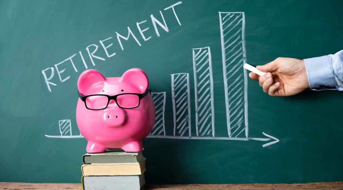 A hand draws an upward-trending graph on a chalkboard with the word 'Retirement' next to a pink piggy bank on a stack of books, symbolizing planning for retirement savings.