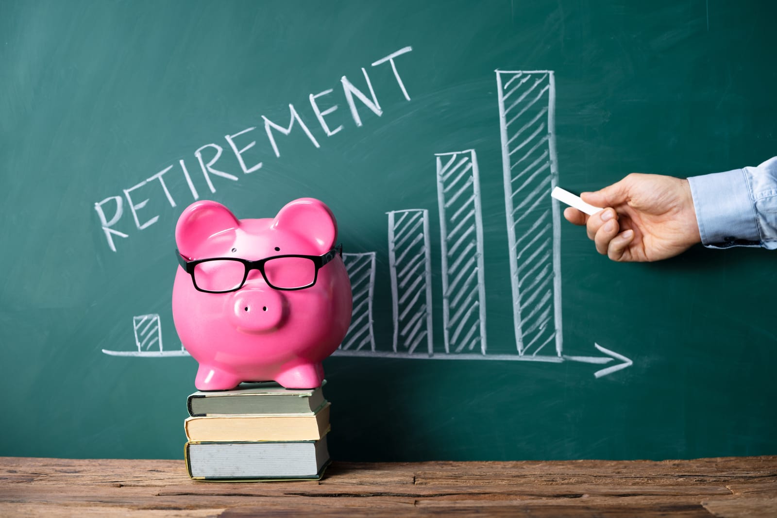 A hand draws an upward-trending graph on a chalkboard with the word 'Retirement' next to a pink piggy bank on a stack of books, symbolizing planning for retirement savings.