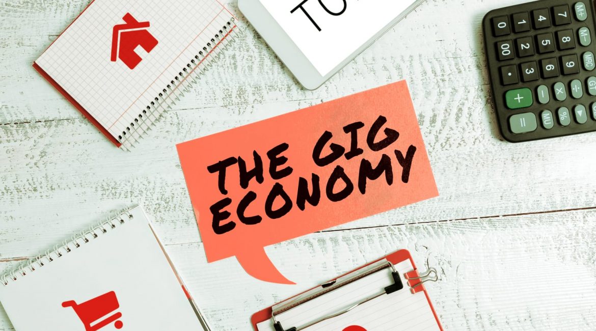 A flat lay image on a white desk with a red speech bubble that says 'THE GIG ECONOMY', surrounded by a calculator, notepads, and a tablet, representing the different facets of gig work.