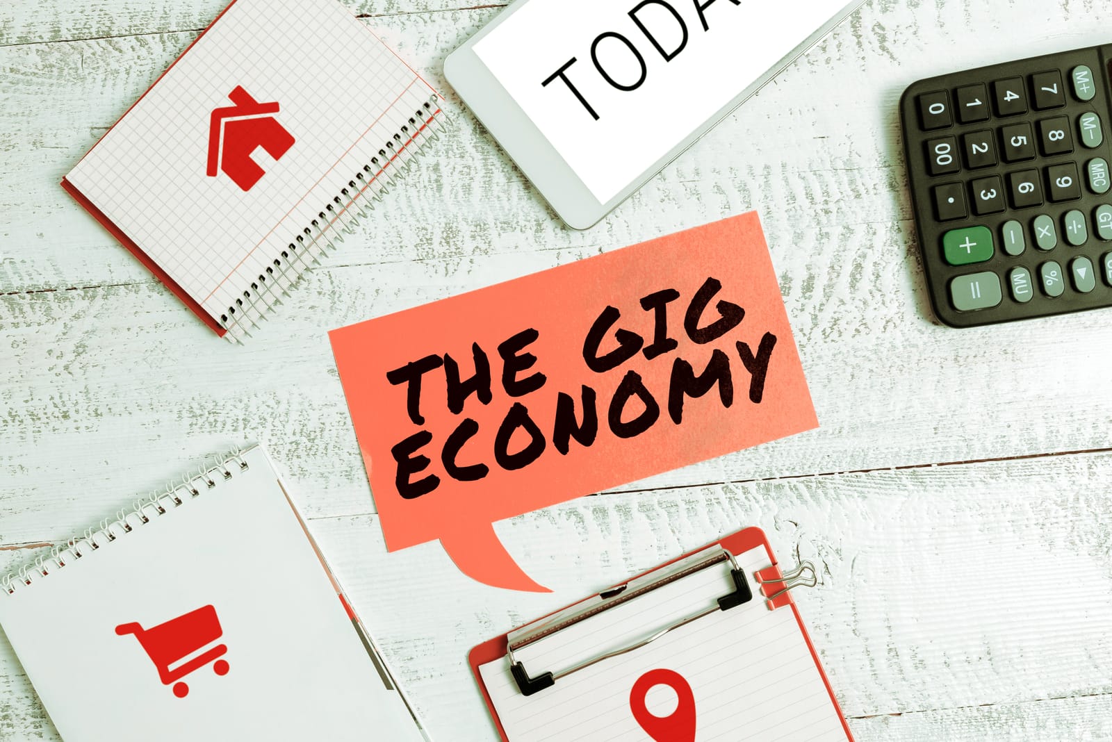 A flat lay image on a white desk with a red speech bubble that says 'THE GIG ECONOMY', surrounded by a calculator, notepads, and a tablet, representing the different facets of gig work.
