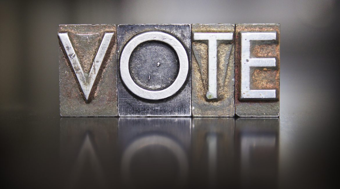 A close-up, artistic shot of old metal letterpress blocks spelling out the word VOTE, representing the topic of elections and electoral reform.